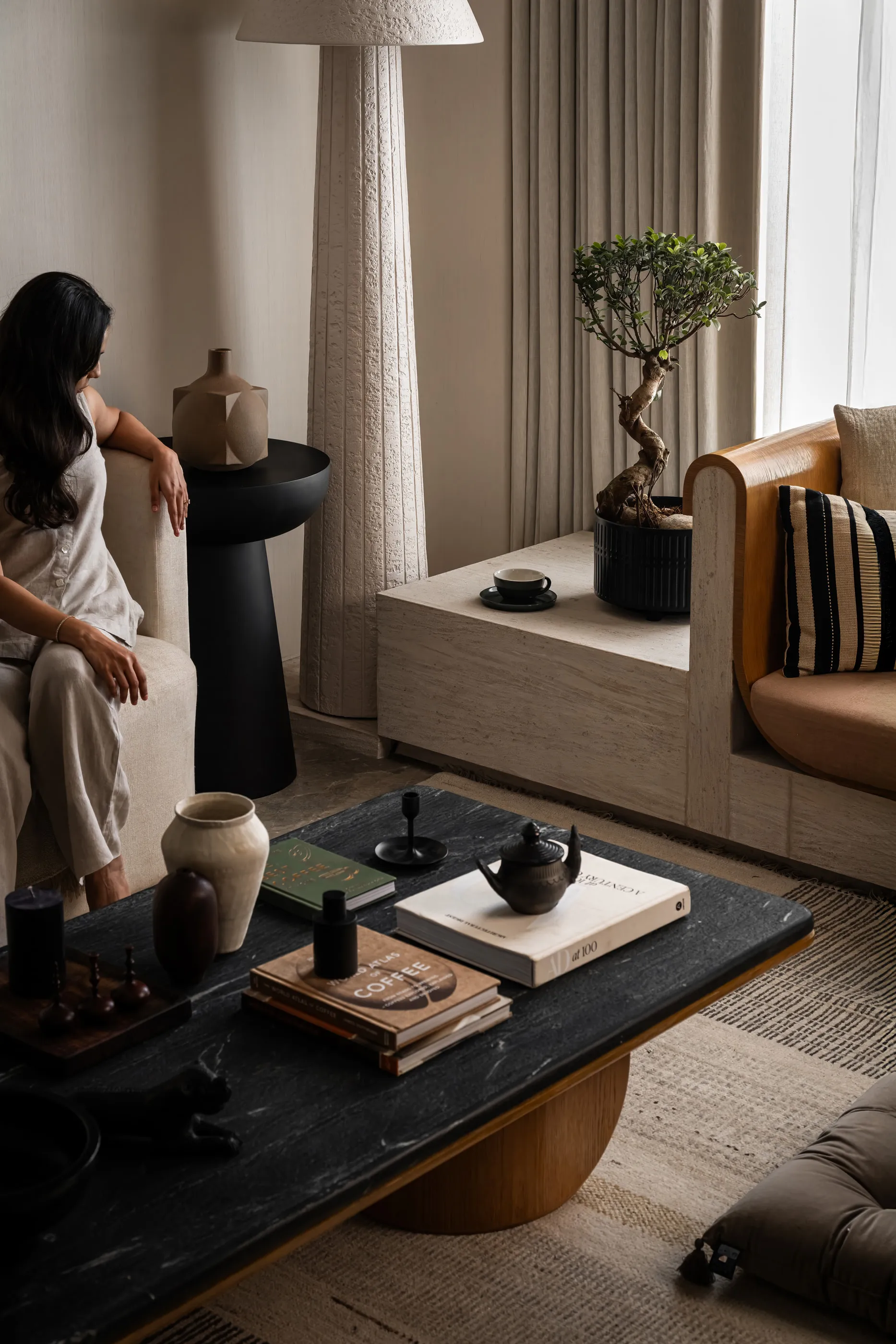 Living-room corner with a textured floor lamp, a bonsai, built-in seating and a black stone coffee table, a woman seated at left