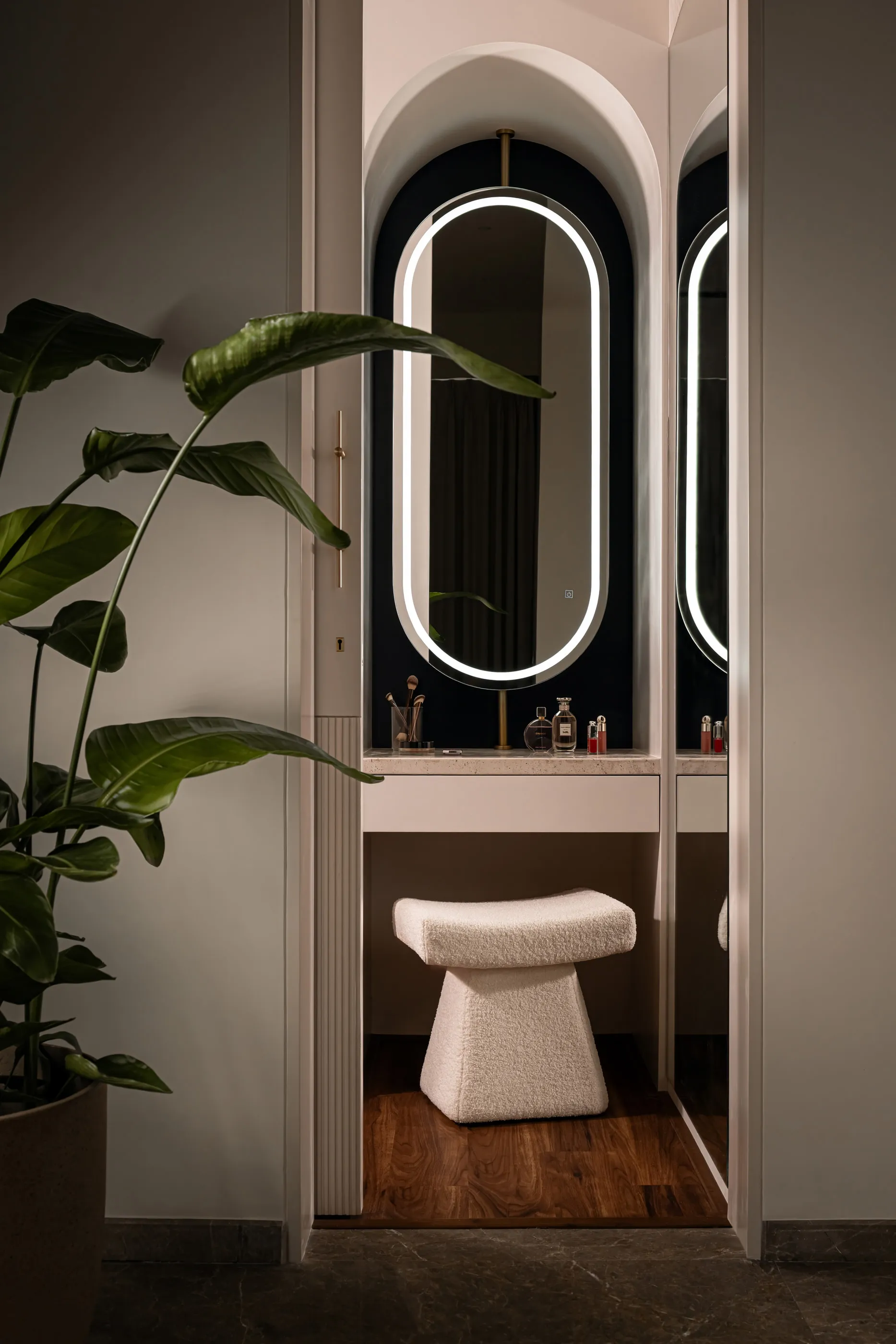 Arched vanity niche with an oval backlit mirror, travertine counter and a boucle stool